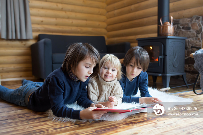 Three sweet children, siblings lying on the floor in little fancy wooden cottage, reading a book, drinking tea