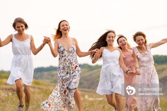 The company of cheerful female friends have a great time together on a picnic in a picturesque place overlooking the green hills. Girls in white dresses dancing in the field