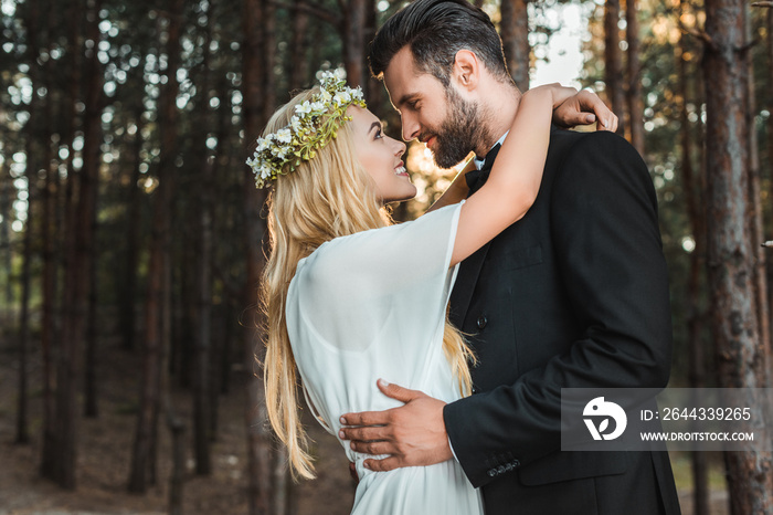 beautiful happy bride in white dress and handsome groom in suit hugging and going to kiss in forest