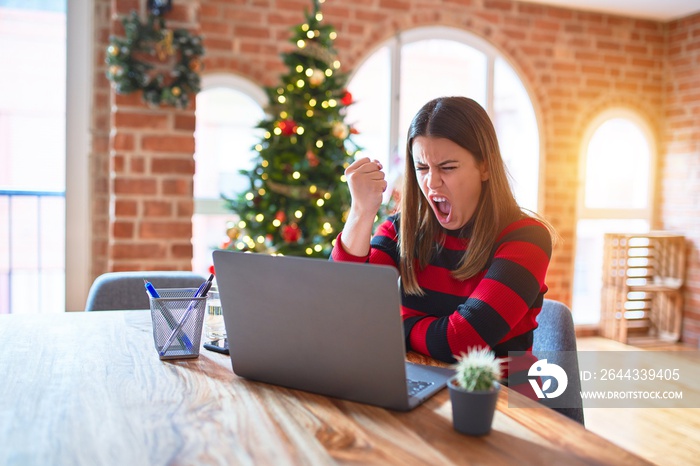 Beautiful woman sitting at the table working with laptop at home around christmas tree angry and mad raising fist frustrated and furious while shouting with anger. Rage and aggressive concept.