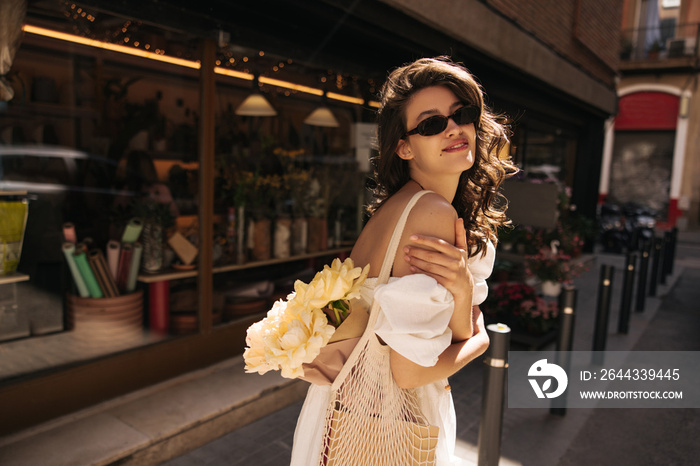 Pretty young caucasian girl posing looking at camera standing outdoors in sunny weather. Brunette woman with wavy hair wears casual summer clothes. Lifestyle concept