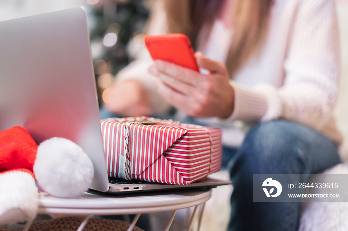 Close up woman with mobile phone, present gift box and laptop. Online shopping at Christmas holidays. Girl having a video chat at home and texting message. Merry Christmas and happy new year.