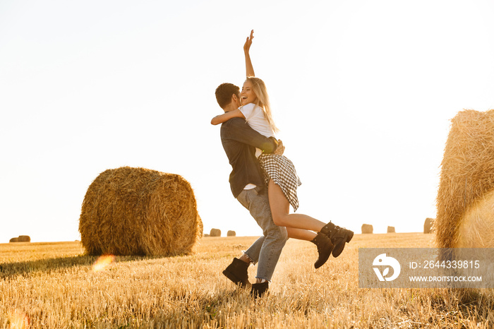 Photo of young couple man and woman having fun while walking through golden field with bunch of haystacks, during sunny day