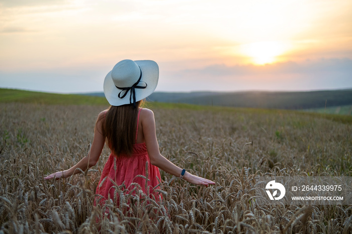 Young happy woman in red summer dress and white straw hat standing on yellow farm field with ripe golden wheat enjoying warm evening.