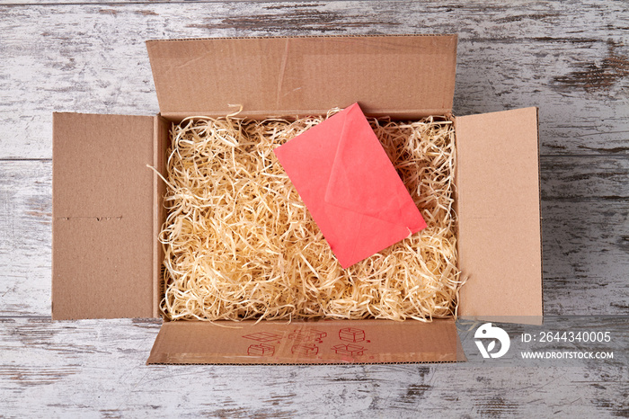 An open parcel with filler. Pink envelope in cardboard box on wooden floor.