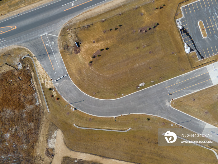 Birds eye view of road intersection with various markings. Fencing along the road. Partial parking lot scene with markings. Open dry grass all around with a free trees. Utility poles with shadows.