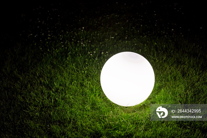 illuminating conglobate ground garden lamp of white color lies on a green lawn in grass in the backyard of park, closeup of lighting fixture night scene in dark outdoor.