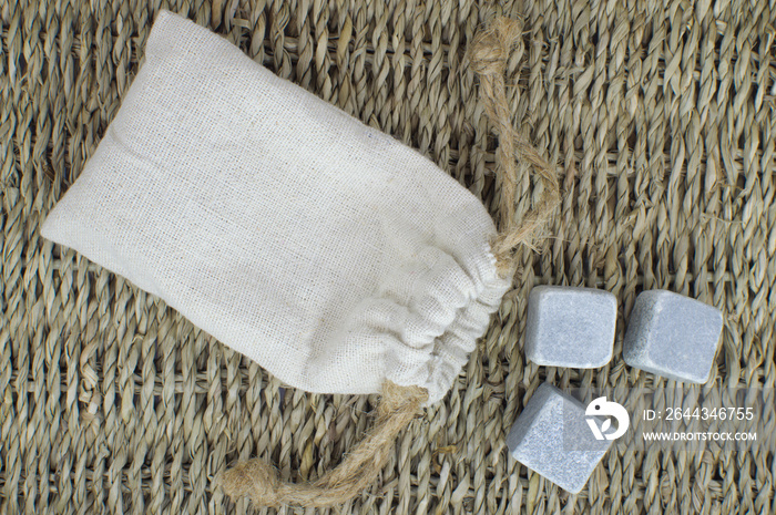 Small eco-bag made of natural linen fabric with rope ties and stones for whiskey cooling. Rough canvas bag and marble cubes on the background of a wicker table. Set for an alcoholic drink.