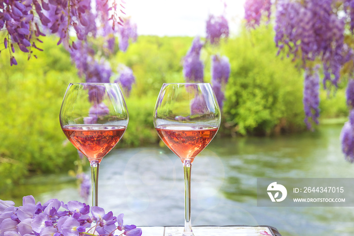 Two glasses of pink wine on a table in front of the river and blooming purple wisteria