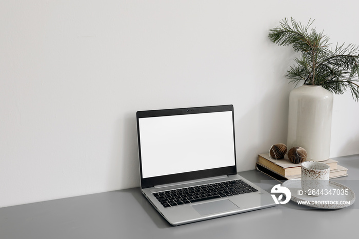 Christmas home office still life. Laptop, computer mockup. Blank screen. Cup of coffee, pine tree branches in vase with Christmas ornaments on grey table. Scandinavian festive winter home decor.