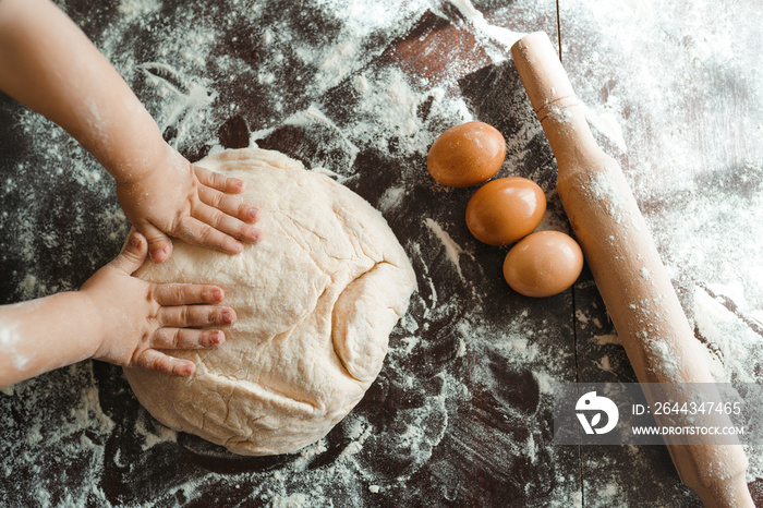 Small hands kneading dough. Little child preparing dough for backing. Kid’s hands, some flour, wheat dough and rolling pin on the dark table.Cooking concept.