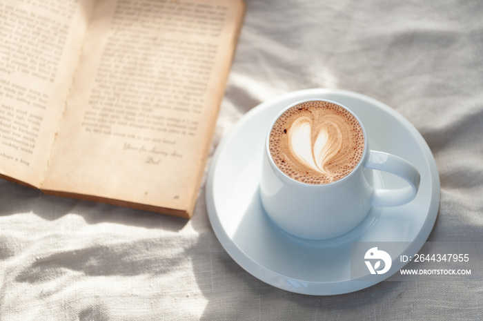 Relaxing holiday morning with hot heart-shaped latte art coffee in a white coffee mug put on the book with the warm morning sunshine from the window to feel relaxed good for relaxing