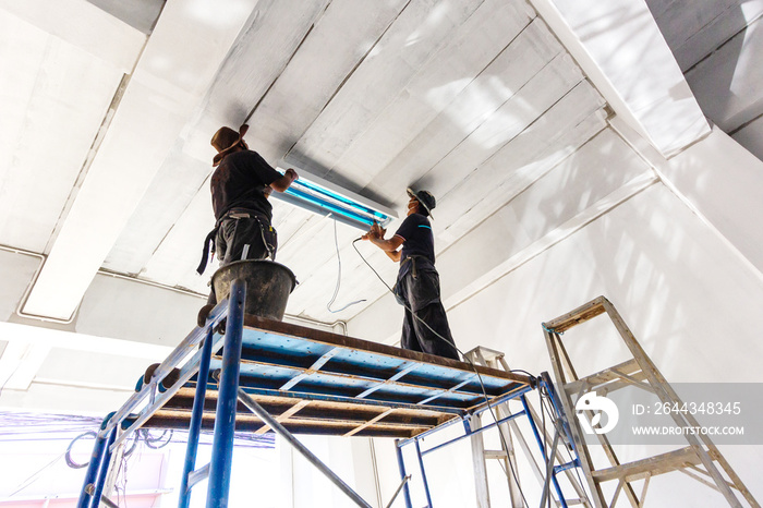 Two technicians standing steel scaffolding is installing fluorescent bulbs on ceiling of building. Home improvement concept.