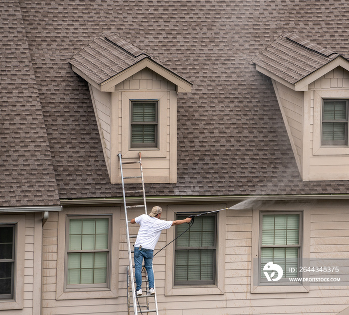 Painter on ladder using pressure wash spray to clean woodwork before painting townhome