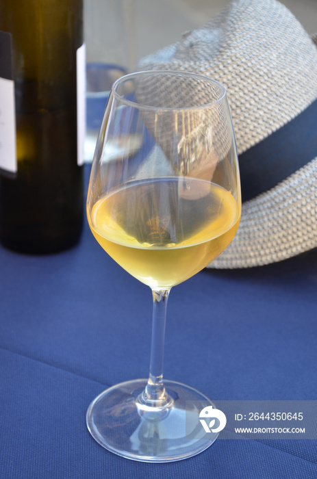 White wine in a crystal glass on a blue covered table, wine bottle and straw hat behind, outdoor in Tuscany, Italy