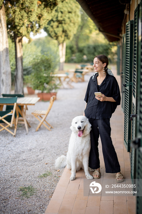 Young caucasian woman standing with Maremmano-Abruzzese Sheepdog and looking away near entrance to hotel room. Concept of weekend, rest and vacation. Smiling beautiful girl with smartphone