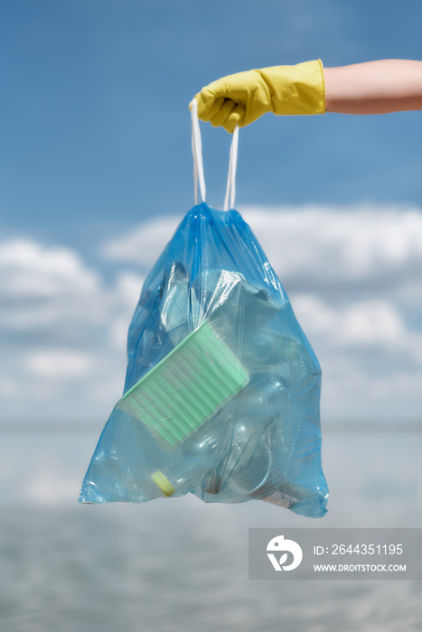 Vertical shot of eco activist wearing yellow rubber gloves holding trash bag with plastic waste and other garbage against sea and sky while cleaning the beach