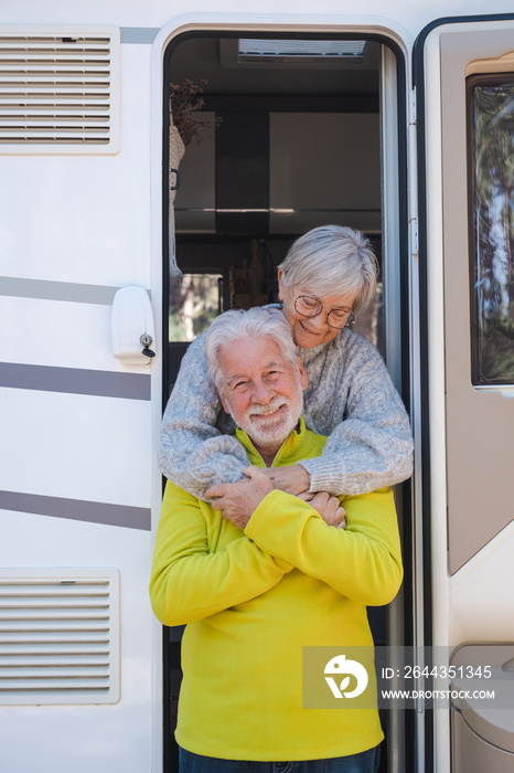 Happy relaxed caucasian senior couple embracing on the door of their camper van motor home. Smiling attractive elderly people enjoying freedom vacation travel in the mountain.