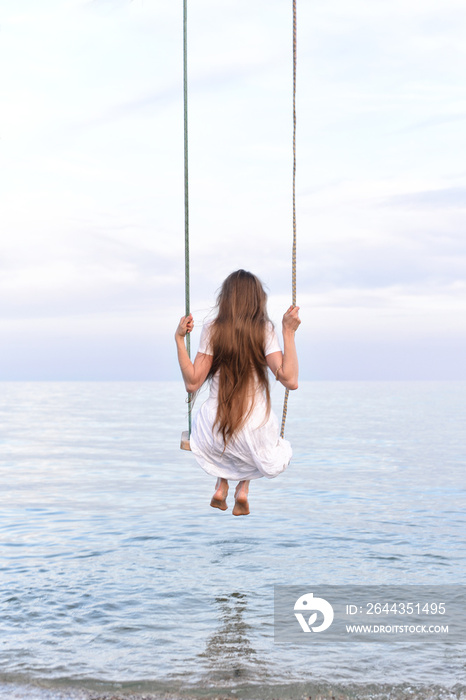 Girl in white dress swinging over the water, enjoying the sea view. Back view