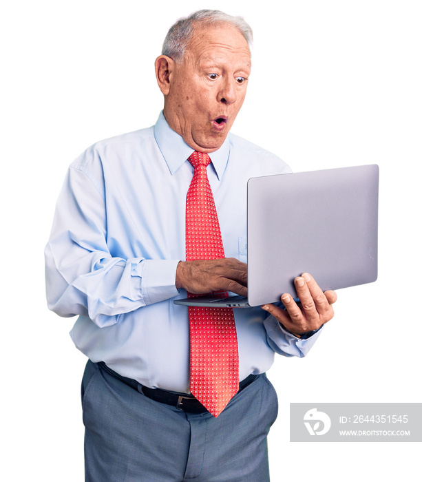 Senior handsome grey-haired man wearing tie using laptop scared and amazed with open mouth for surprise, disbelief face