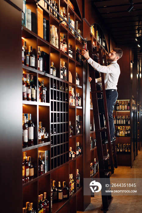 The seller of alcoholic beverages sorts bottles standing on the ladder.