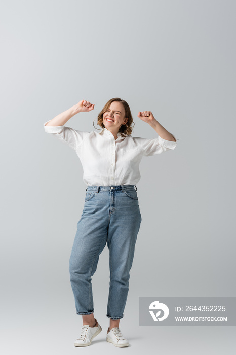 full length of pleased overweight woman in jeans celebrating on grey