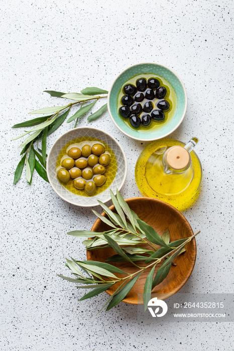 Green and black olives with olive oil in a glass bottle, olive tree sprigs and cut fresh ciabatta bread on wooden cutting board. White rustic background, mediterranean food concept, top view