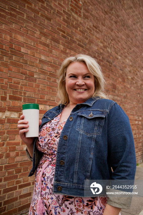 Plus-sized woman in front of brick wall holding a reusable coffee cup and smiling