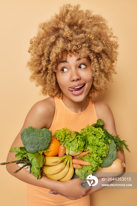 Vertical shot of funny positive curly haired woman sticks out tongue looks away poses with bih heap of freshly picked vegetables and fruits eats healthy organic food isolated over beige background