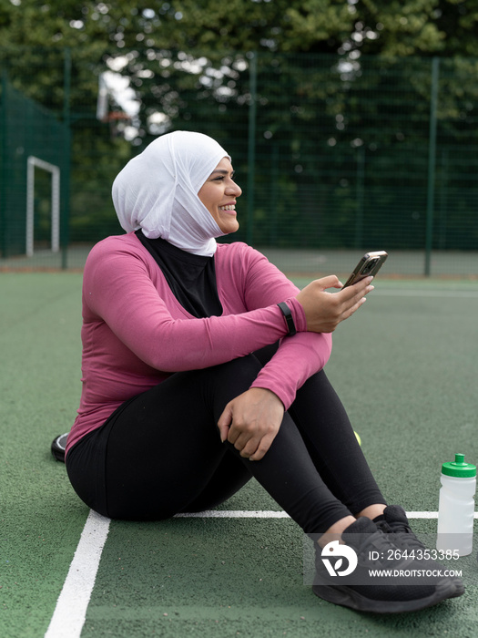 UK,Sutton,Woman in headscarf sitting on tennis court,holding smart phone