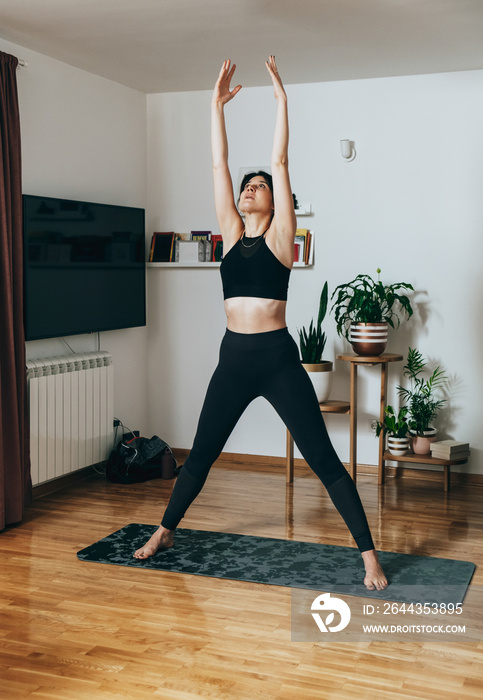 Beautiful fitness woman in black sportswear stretching arms and shoulders and doing breathing exercises while standing on a gray exercise mat in the living room at home