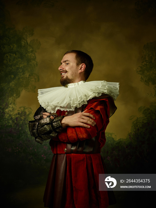 Young man as a medieval knight on dark studio background. Portrait in low key of male model in retro costume. Standing and smiling. Human emotions, comparison of eras and facial expressions concept.