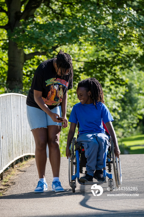 Teenage girl (16-17) in wheelchair with friend walking in park