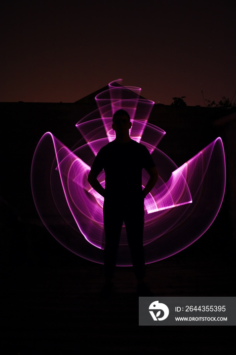 Silhouette of a man standing from the front. Curved abstract shapes made of violet light saber in background. Lightpainting session in long exposure at night.