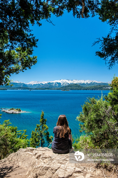 Young woman sitting on the rock and watching at Nahuel Huapi lake, San Carlos de Bariloche Argentina. Waves on the lake. Mountains with fresh snow surrounding the lake.