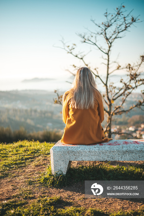 Woman on a yellow coat sitting on a bench contemplating the landscape