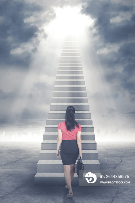 Businesswoman walking up staircase to door in sky with bright light shining down