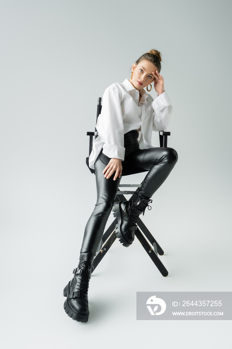 full length of stylish woman in white shirt and black pants with leather boots sitting on chair on grey background.