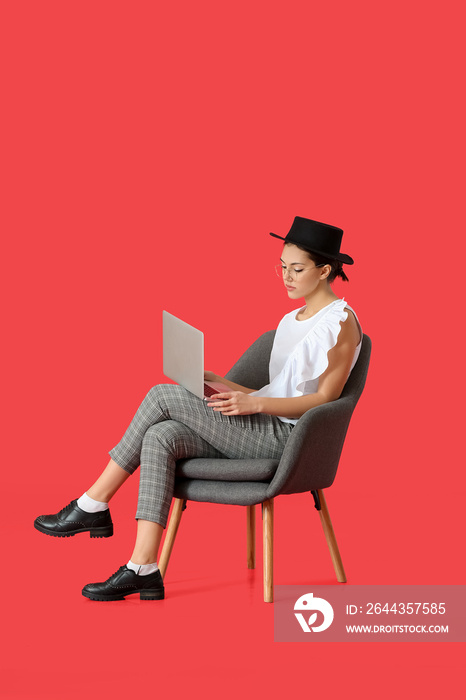 Young woman with laptop sitting in armchair on color background