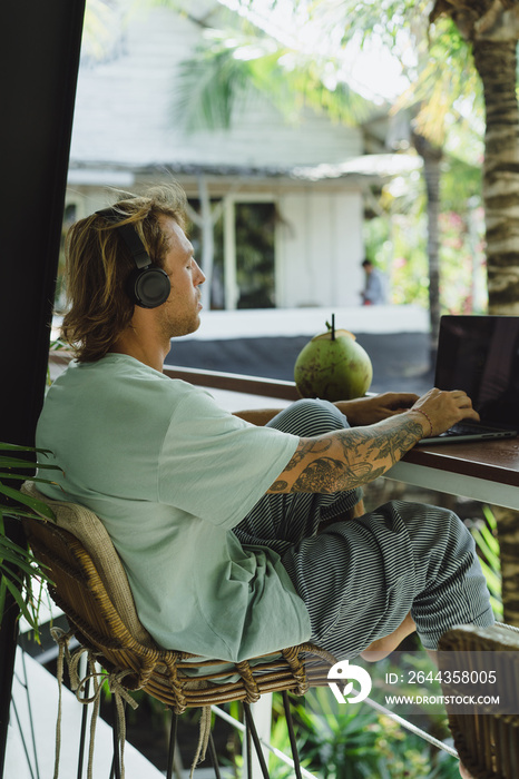 A young smiling man in big headphones works on a laptop in a Bali coworking space. Daily call with the team. Work on your laptop from anywhere.