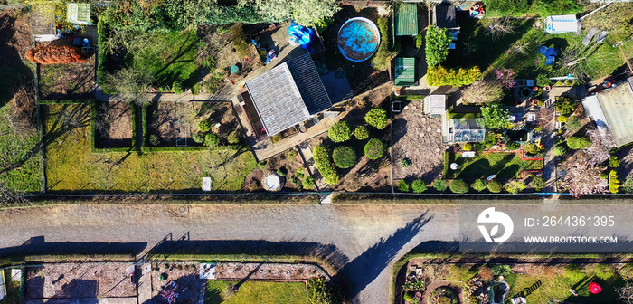 Abstract vertical aerial view of garden arbor in allotment garden behind dead straight gray path with ornamental trees and useful plants