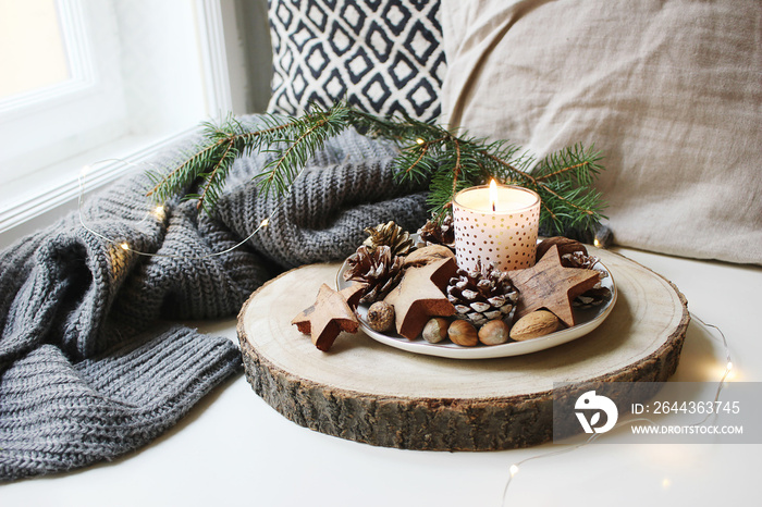 Winter festive still life scene. Burning candle decorated by wooden stars, hazelnuts and pine cones standing near window on wooden cut board. Glittering Christmas lights. Fir branch on wool plaid.