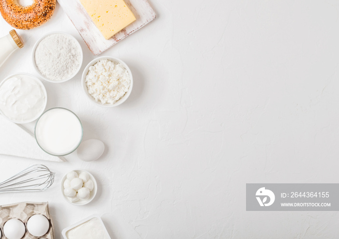 Fresh dairy products on white table background. Glass of milk, bowl of sour cream and cottage cheese and eggs. Fresh baked bagel. Steel whisk. Top view.Space for text