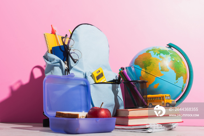 School backpack full of stationery near lunch box, globe, school bus model and books on pink