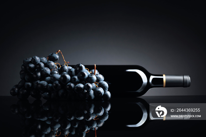 Bottle of red wine and a bunch of grapes on a black reflective background.