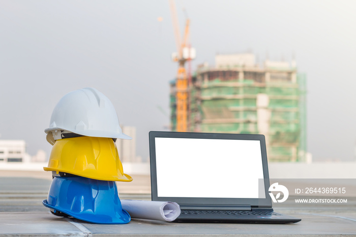 The safety helmet and the blueprint with laptop has white screen isolated at construction site with crane background