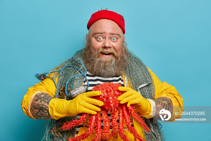 Image of happy positive lobsterman glad to catch big octopus, enjoys fishing at sea, has lucky day, wears red hat, yellow gloves, poses over blue background. Bearded sailor has marine travel