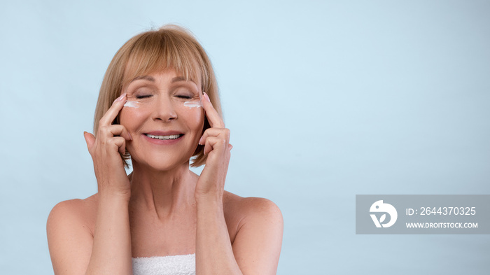 Mature beauty concept. Lovely senior woman applying face cream under her eyes over blue background, banner design