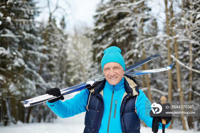 Happy senior sportsman with skis looking at you while standing in forest on winter day