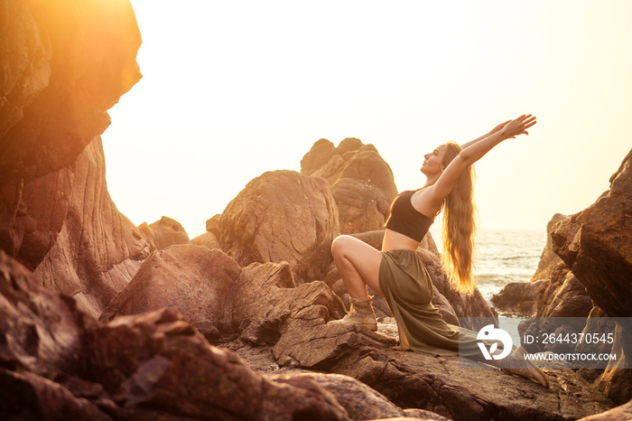 stylish redheaded chestnut blonde young girl in indie bohemian bo-ho style clothes black stylish top.boho woman long hair,makeup and green feathers in head practicing yoga asanas on the rocks by sea